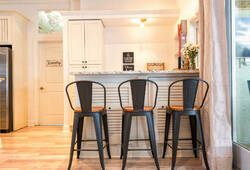 Stylish metal bar stools invite you to gather around the sleek granite countertop, while soft natural light filters through the large windows, illuminating the modern kitchen's subtle coastal decor and pristine cabinetry.