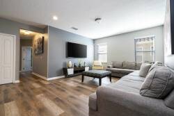 This bright living room boasts a spacious gray sectional and a sleek coffee table, perfect for relaxing. Large windows fill the space with natural light, highlighting the warm wood tones of the flooring for a cozy ambiance.