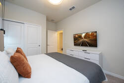 This cozy bedroom features a plush bed adorned with soft gray and warm orange pillows, inviting you to unwind. A sleek white dresser complements the space, while a mounted TV offers entertainment at your fingertips.