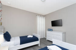 This inviting bedroom features two cozy twin beds with crisp white linens and deep blue accents for a refreshing vibe. Natural light streams through sheer curtains, while a sleek dresser and wall-mounted TV enhance your comfort.