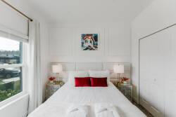 This bright bedroom, featuring a plush white bed with vibrant red pillows, invites restful sleep. Stylish bedside tables and lamps create a warm ambiance, while large windows offer lovely views and abundant natural light.