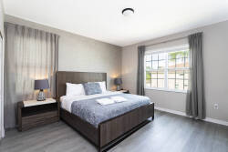 This spacious bedroom boasts a stylish wooden bed frame with plush bedding for a restful atmosphere. Soft gray tones enhance the natural light from the large window, and elegant curtains provide an inviting touch of warmth.