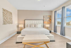 Rest in a bright and elegant bedroom with a plush upholstered bed, soft bedding, stylish bench, bedside lamps, wall art, large windows, and warm neutral tones for a relaxing stay.
