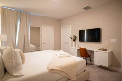 This cozy bedroom, adorned in soft, neutral tones with plush bedding, features a stylish throw and a sleek desk area, perfect for morning coffee. Natural light filters through elegant curtains, enhancing the tranquil ambiance.