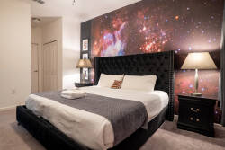 Relax in a galactic space-themed bedroom with a bold starry mural, elegant tufted headboard, cozy bedding, and dark accent furniture. Soft lighting, framed character art, and gray tones create a fun and comfortable retreat for vacation.