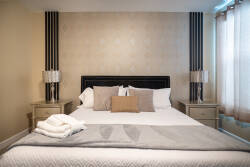 This beautifully styled bedroom features a plush bed with soft pillows and a cozy throw, paired with elegant lamps for a warm ambiance. Subtle wall tones and natural light from sheer curtains enhance the inviting atmosphere.