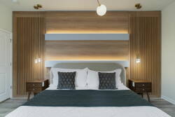 Experience restful nights in this beautifully designed bedroom, featuring a plush bed adorned with textured pillows and a deep green throw. Warm wood paneling and stylish bedside lamps create an inviting atmosphere for relaxation.