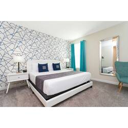 Rest in a bright modern bedroom with a comfortable bed, white bedding, blue accent pillows, geometric wall design, teal curtains, bedside lamps, soft carpet, and a stylish accent chair.