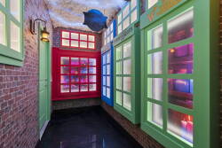 Enter a vibrant corridor with colorful windows framing natural light that dances across the polished black floor. The rustic charm of exposed brick walls creates a cozy and playful atmosphere, inviting you to explore.