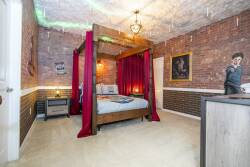 Step into a unique retreat featuring a striking four-poster bed draped in rich red fabric, complemented by exposed brick walls and soft carpeting. The warm glow of the chandelier adds a cozy atmosphere, perfect for relaxation.