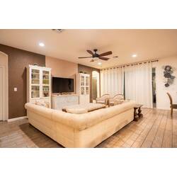 Relax in a warm and elegant living room with a spacious sectional sofa, wall-mounted TV, white cabinets, and soft natural light through the curtains. Neutral tones, wood floors, and cozy seating create a welcoming space for family time.