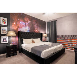 Relax in a galactic space-themed bedroom with a bold starry mural, elegant tufted headboard, cozy bedding, and dark accent furniture. Soft lighting, framed character art, and gray tones create a fun and comfortable retreat for vacation.