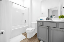 Enjoy this modern bathroom featuring bright white walls, sleek grey cabinetry, and warm wood-like flooring bathed in natural light. Relax in the spacious tub and appreciate the thoughtfully designed vanity for your personal care.