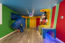 Step into a vibrant playroom designed for fun, featuring bold, colorful walls and a playful slide. Mickey Mouse adds a whimsical touch, while the soft flooring ensures safety and comfort for kids of all ages.