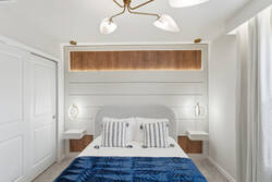 Rest in a bright modern bedroom with a cozy white bed, blue throw, wood accent wall, soft pendant lights, white curtains, wall-mounted TV, and calm neutral details for vacation comfort.