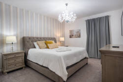 Enjoy a tranquil bedroom adorned in soft gray hues and elegant striped wallpaper. A cozy tufted bed awaits, illuminated by a warm chandelier, while decorative pillows invite you to relax and unwind in comfort.