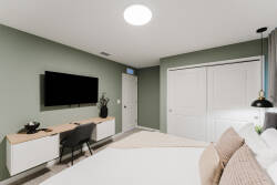 This thoughtfully designed bedroom features calming green walls, a cozy bed with soft pillows, a sleek desk, and a modern TV, all bathed in natural light that enhances its inviting atmosphere.