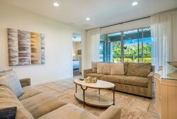 Unwind in the cozy living area with a plush beige sectional and a stylish round coffee table perfect for conversations. Large windows with sheer curtains fill the room with natural light, showcasing the beautiful outdoor scenery.