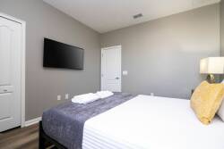 This cozy bedroom boasts a sleek gray accent wall and a comfy bed with crisp white linens and a textured throw. A mounted flat-screen TV adds entertainment, while natural light pours in, creating a warm and inviting atmosphere.
