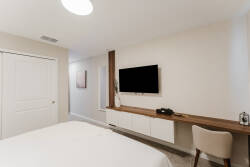 Enjoy a cozy bedroom with a plush bed, soft linens, a sleek wooden desk, and a modern wall-mounted TV, all set in neutral tones and bathed in natural light for a warm, inviting atmosphere during your stay.