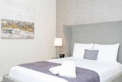 Experience a cozy bedroom with a plush gray headboard, crisp white linens, and a soft purple throw. Enjoy warm lighting from a stylish lamp and the calming ambiance of a serene landscape painting in your restful retreat.