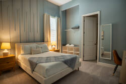 Basked in soft natural light, this cozy bedroom features calming blue walls and plush carpet, creating a tranquil atmosphere. The stylish furnishings, including a sleek mirror and warm wood accents, enhance comfort and relaxation.