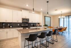 Cook and gather in a bright modern kitchen with white cabinets, stainless steel appliances, a large island, pendant lights, black bar seating, and a dark tile backsplash, perfect for family meals during vacation.