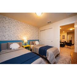 Rest in a cozy twin bedroom with brick-style accent wall, blue headboard, navy throws, yellow nightstand, soft lamp, wall-mounted TV, and playful decor for vacation comfort.