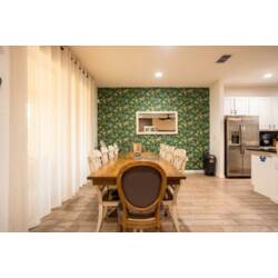 Share meals in a bright dining area with a long wood table, comfortable seating, soft curtains, and a leafy accent wall with a mirror. The nearby kitchen and warm lighting make this space welcoming for family meals.