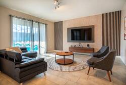 Relax in a modern living room with a black leather sofa, gray accent chairs, wall-mounted TV, wood slat details, soft curtains, pool views, and a round coffee table, creating a comfortable space for vacation moments.