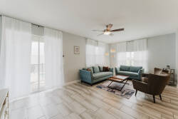 This cozy living room features soft, teal couches that invite you to unwind while overhead, a stylish ceiling fan circulates a gentle breeze. Natural light pours in through sheer curtains, enhancing the warm, welcoming ambiance.