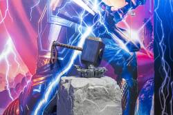 Discover a vibrant space alive with dynamic art, showcasing a stunning mural of a powerful hero in electric blues and purples. At the center, a replica of a legendary hammer on a textured stone pedestal sparks your sense of.