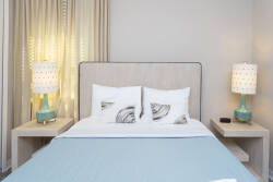 Experience a cozy bedroom with a plush bed dressed in crisp white linens and a soothing blue throw, stylish turquoise lamps on chic nightstands, and soft natural light gently filtering through elegant drapes for a serene ambiance.