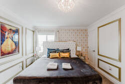 Enter a stylish sanctuary with a plush king bed dressed in rich, golden accents and luxurious linens. Elegant artwork and intricate wall details enhance the room's charm, illuminated by soft natural light from the window.