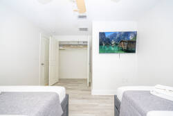 This bright and airy room offers two cozy beds dressed in soft gray linens, a sleek wall-mounted TV, and light wood flooring that enhances the spacious atmosphere, plus a closet for convenient storage of your belongings.