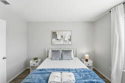 Rest in a bright bedroom with a cozy white bed, soft blue throw, gray pillows, abstract wall art, natural light, and white curtains, creating a calm space to enjoy your vacation.