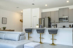 The modern kitchen features stylish grey cabinetry and gold-accented bar stools, perfect for casual meals or morning coffee. Abundant natural light fills the open layout, creating an inviting atmosphere for your gatherings.