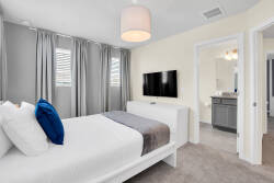 Experience a serene bedroom with soft gray curtains that let in natural light. Relax on the cozy bed with a plush white duvet and vibrant blue accent pillow, complemented by a sleek TV for modern convenience.