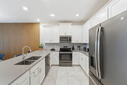 Cook with ease in a bright modern kitchen with white cabinetry, stainless steel appliances, a large island, sleek sink, bright lighting, and warm wood accents nearby.