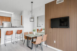 Enjoy a modern dining area that flows into a chic kitchen with warm wooden cabinets and sleek bar seating. The textured accent wall and stylish lighting offer a cozy ambiance perfect for meals or relaxation.