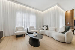 This bright and airy living area features a plush curved sofa and minimalist armchairs, enhanced by soft curtains. The warm wood accents and sleek coffee table provide a stylish yet comfortable space ideal for relaxation.