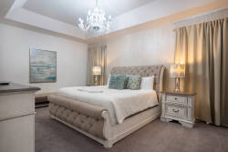 This elegant bedroom boasts a plush, tufted bed with crisp white linens and soothing teal throw pillows. Graceful drapes frame the windows, while a beautiful chandelier adds a warm glow for a cozy evening atmosphere.