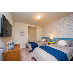Rest in a cozy twin bedroom with brick-style accent wall, blue headboard, navy throws, yellow nightstand, soft lamp, wall-mounted TV, and playful decor for vacation comfort.