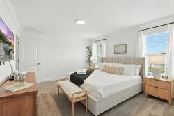 This bright bedroom welcomes natural light through large windows, enhancing the soft bedding and stylish wooden nightstands. A cozy bench at the foot of the bed offers added comfort and functionality for your stay.