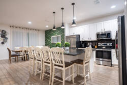 Gather in a bright open kitchen and dining area with white cabinets, stainless steel appliances, black countertops, and island seating. Pendant lights, wood-look floors, and a large dining table create a welcoming space for family meals.