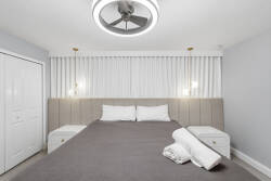 Unwind in this beautifully designed bedroom with a cozy gray bedspread and stylish bedside tables. Soft white curtains and a modern ceiling fan enhance the tranquil atmosphere, perfect for restful nights.