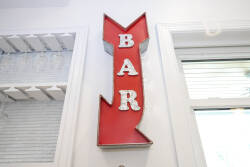 Experience a vibrant atmosphere highlighted by a unique vintage red "BAR" sign. Bright white walls and natural light contribute to an energetic vibe, ideal for unwinding with friends or savoring a casual drink.