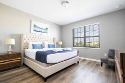 This beautifully designed bedroom invites relaxation with its plush tufted headboard and crisp white linens. Natural light fills the space through a large window, enhancing the warm wooden floors and soothing neutral tones.