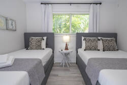 This cozy room offers two twin beds with soft gray linens and stylish patterned pillows, creating a relaxed atmosphere. Natural light floods in through the window, enhancing the modern decor and inviting a refreshing ambiance.