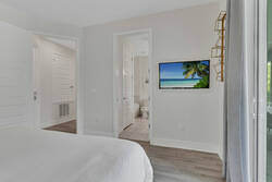 Rest in a bright bedroom with soft white bedding, a wall-mounted TV, light wood flooring, gray curtains, sliding glass doors, and direct access to a private bathroom for extra comfort.